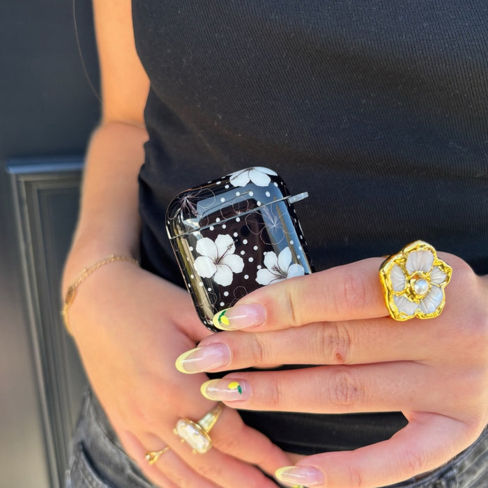 Hand holding the Coconut Lane Mono Flora AirPods case with black top, gold jewelry, and a floral ring visible.