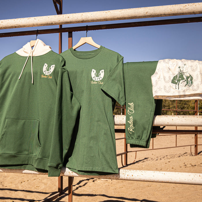 Green 'Rodeo Club' apparel hanging on a wooden fence with a clear blue sky.