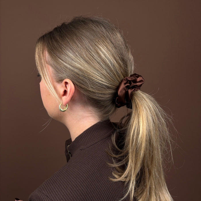 Blonde woman with ponytail wears a brown Coconut Lane scrunchie and dark top, facing away from the camera.