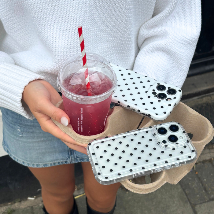 Person in white sweater holds two clear phone cases with black polka dots and a pink drink.
