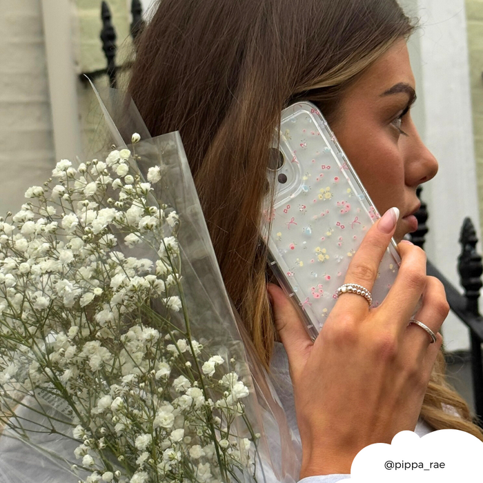 Woman holding a clear Coconut Lane Bows in Bloom phone case with floral accents to her ear, facing sideways outdoors.