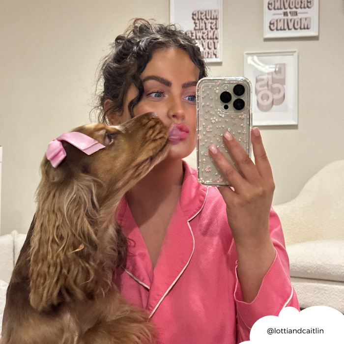 Woman in pink pajamas takes a mirror selfie with a clear phone case featuring bow patterns; her dog licks her face.