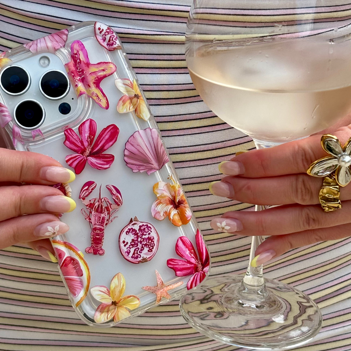 Hand holding clear Rosé Coast phone case with pink accents and a glass of white wine; manicured nails visible.
