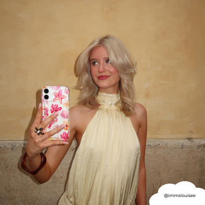 Blonde woman in a cream halter dress holds a clear phone case with pink and gold accents, taking a selfie.
