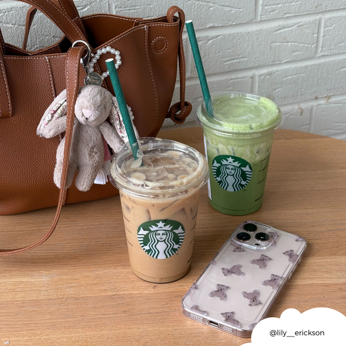 Clear Coconut Lane phone case, brown bag, and plush teddy keychain next to two Starbucks drinks on a table.