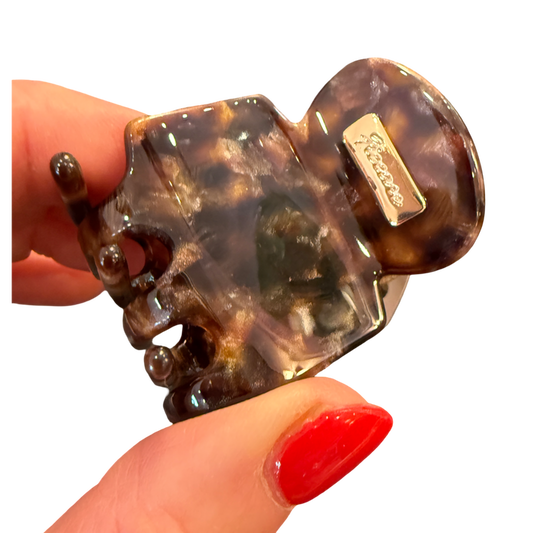 A tortoiseshell hair claw clip held by a hand with red nail polish against a plain white background. The brand name 'Sartoso' is visible on a small metal tag attached to the clip.