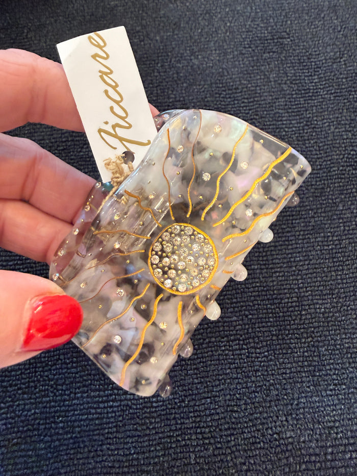 A hand with red nail polish holds a resin hair claw clip featuring a sunburst design with gold lines, a crystal-embellished center, and small pearl-like accents. A white paper tag with the brand name 'Riccare' is attached.
