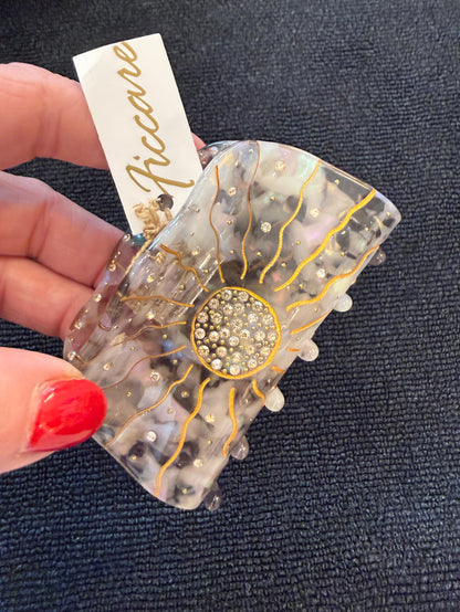 A hand with red nail polish holds a resin hair claw clip featuring a sunburst design with gold lines, a crystal-embellished center, and small pearl-like accents. A white paper tag with the brand name 'Riccare' is attached.