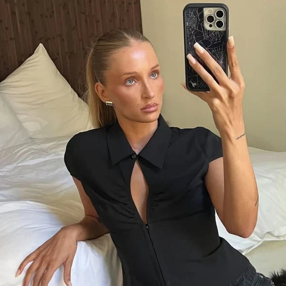 Woman taking a mirror selfie wearing a black sleeveless top with a plunging neckline.