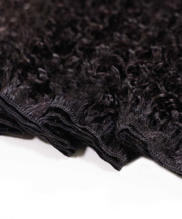 Close-up of several bundles of curly, virgin dark brown or black hair extensions with visible stitching on the wefts, on white surface.