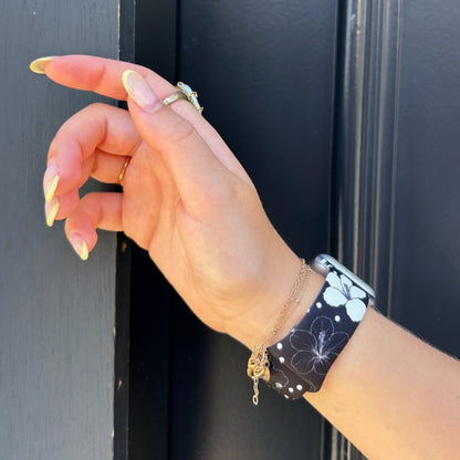 Yellow-manicured hand wearing rings, a gold bracelet, and a Mono Flora patterned Apple Watch strap by Coconut Lane.