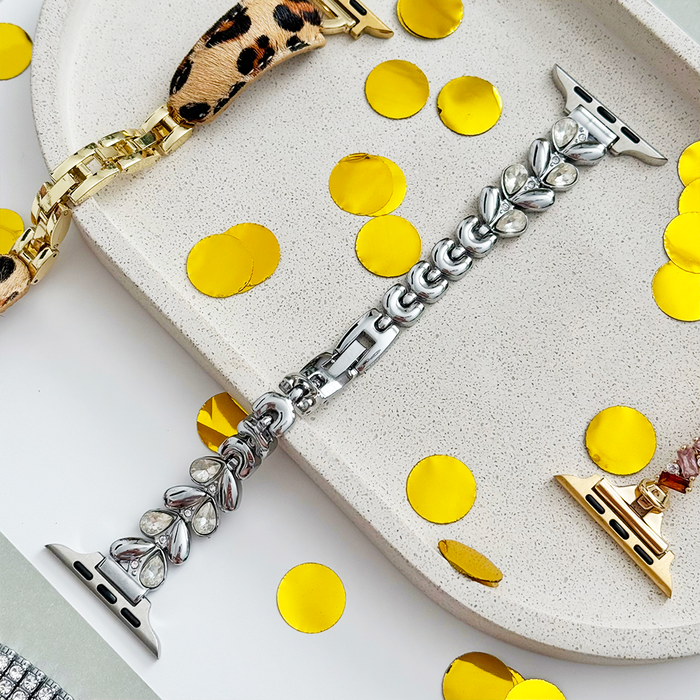 Silver Coconut Lane Sparkle Petal Apple Watch strap on a stone tray, with gold confetti dots and a leopard print band nearby.