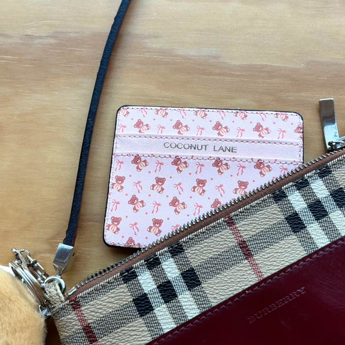 Beige teddy card holder partially visible behind a Burberry-style checked wallet on a light wooden surface.