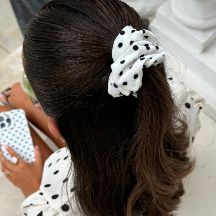 Woman with brown hair wears a white polka dot scrunchie and matching shirt, holding a polka dot phone case.
